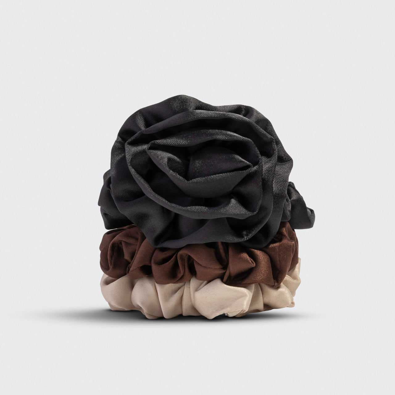 A black rosette scrunchie stacked on 2 cream and brown scrunchies on a light gray background