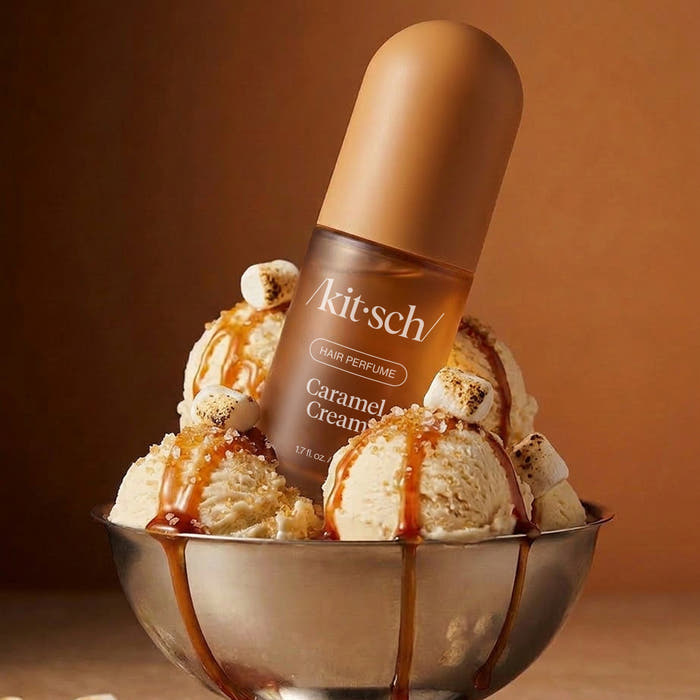 A Kit-sch Caramel Cream perfume bottle stands upright in a metal bowl filled with vanilla ice cream, caramel drizzle, and toasted marshmallows, against a warm brown studio background.