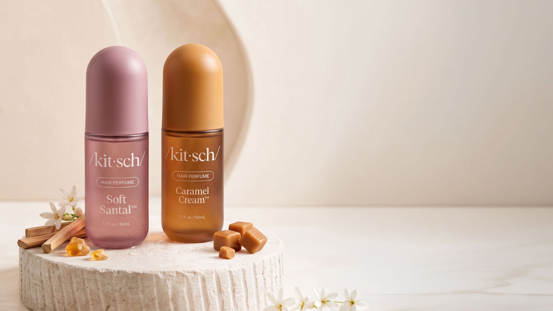 Two Kitsch hair perfume bottles, one in Soft Santal (dusty mauve) and one in Caramel Cream (caramel brown), displayed on a textured stone pedestal with decorative cinnamon sticks and caramel pieces against a warm beige background.